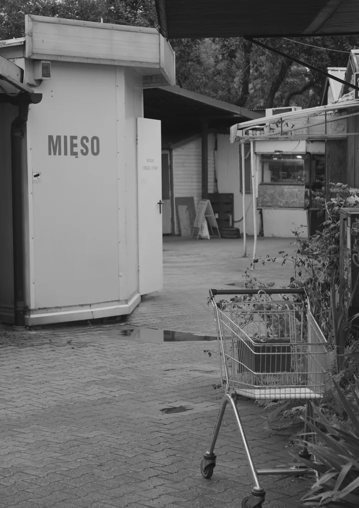 Czarno-biała fotografia przedstawia wejście do pawilonu bazarowego z napisem "MIĘSO". W pierwszym planie widoczny jest wózek sklepowy, a w tle charakterystyczna architektura handlowa. Zdjęcie dokumentuje przestrzeń Bazarku pod Halą Marymoncką, gdzie odbył się koncert Pawła Szamburskiego "Oda do bazarku", dedykowany sprzedawcom bazarowym.