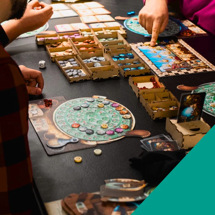 Zdjęcie przedstawia planszę gry strategicznej z sześciokątnymi polami w odcieniach zieleni i brązu, z rozmieszczonymi na niej pionkami i żetonami. Na planszy widoczne są różne elementy rozgrywki, w tym figurki wojskowe i znaczniki. Obrazek promujący cykl "Planszownia" - spotkania przy grach planszowych organizowane regularnie przez Żoliborski Dom Kultury. 